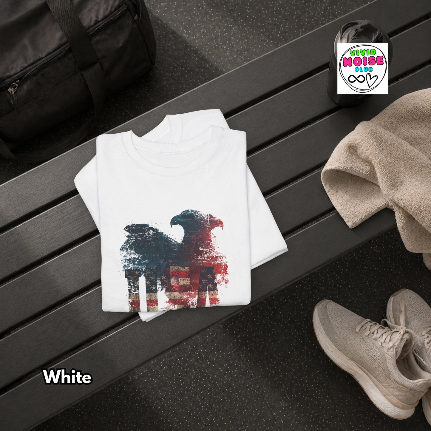 White t-shirt with a graphic design on a black surface, next to a black bag and beige shoes.