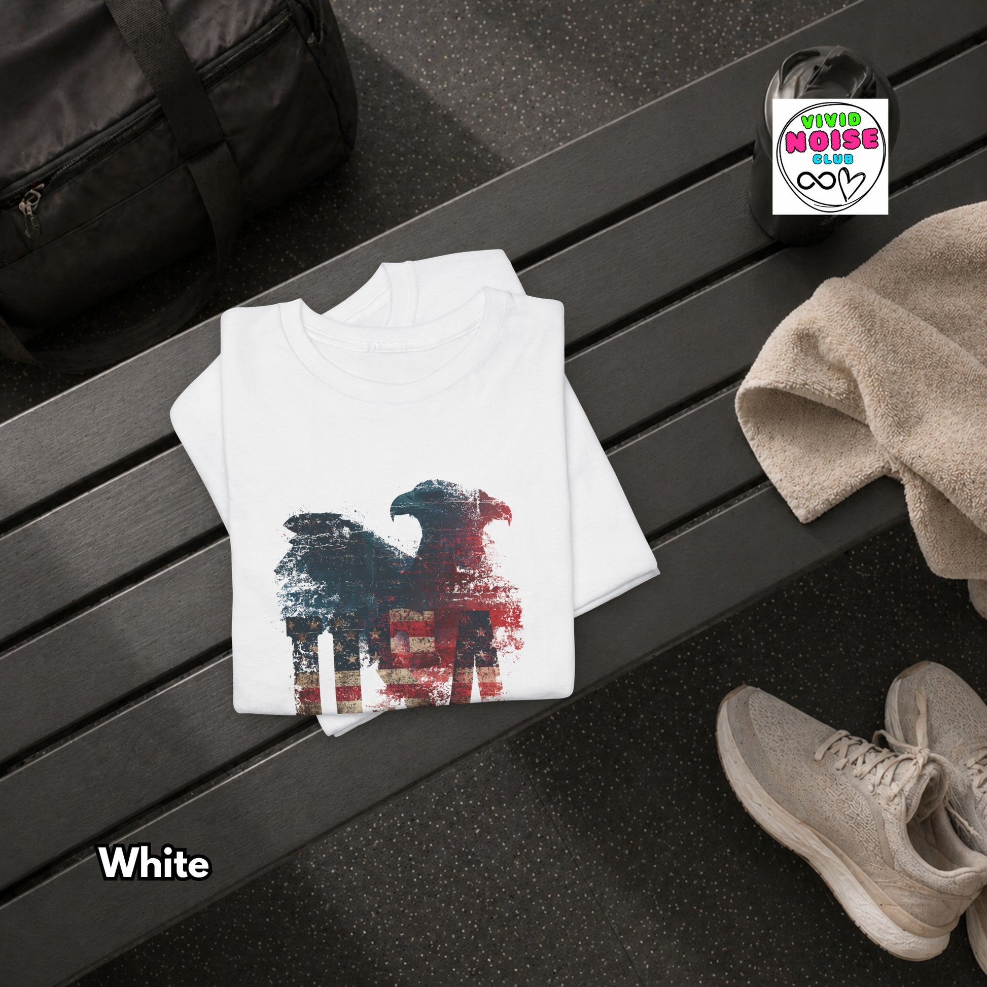 White t-shirt with a graphic design on a black surface, next to a black bag and beige shoes.