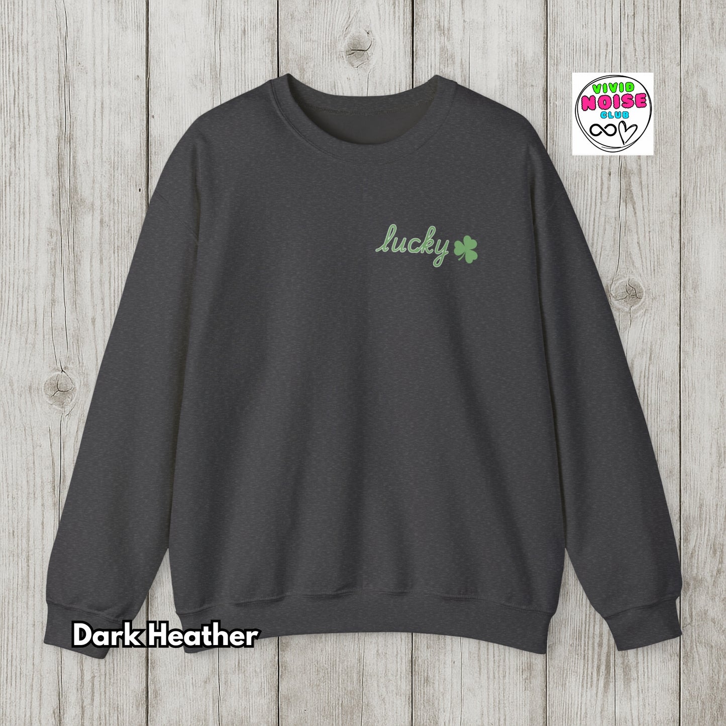 Dark Heather sweatshirt with 'lucky' and clover design on a wooden surface, featuring Vivid Noise logo.