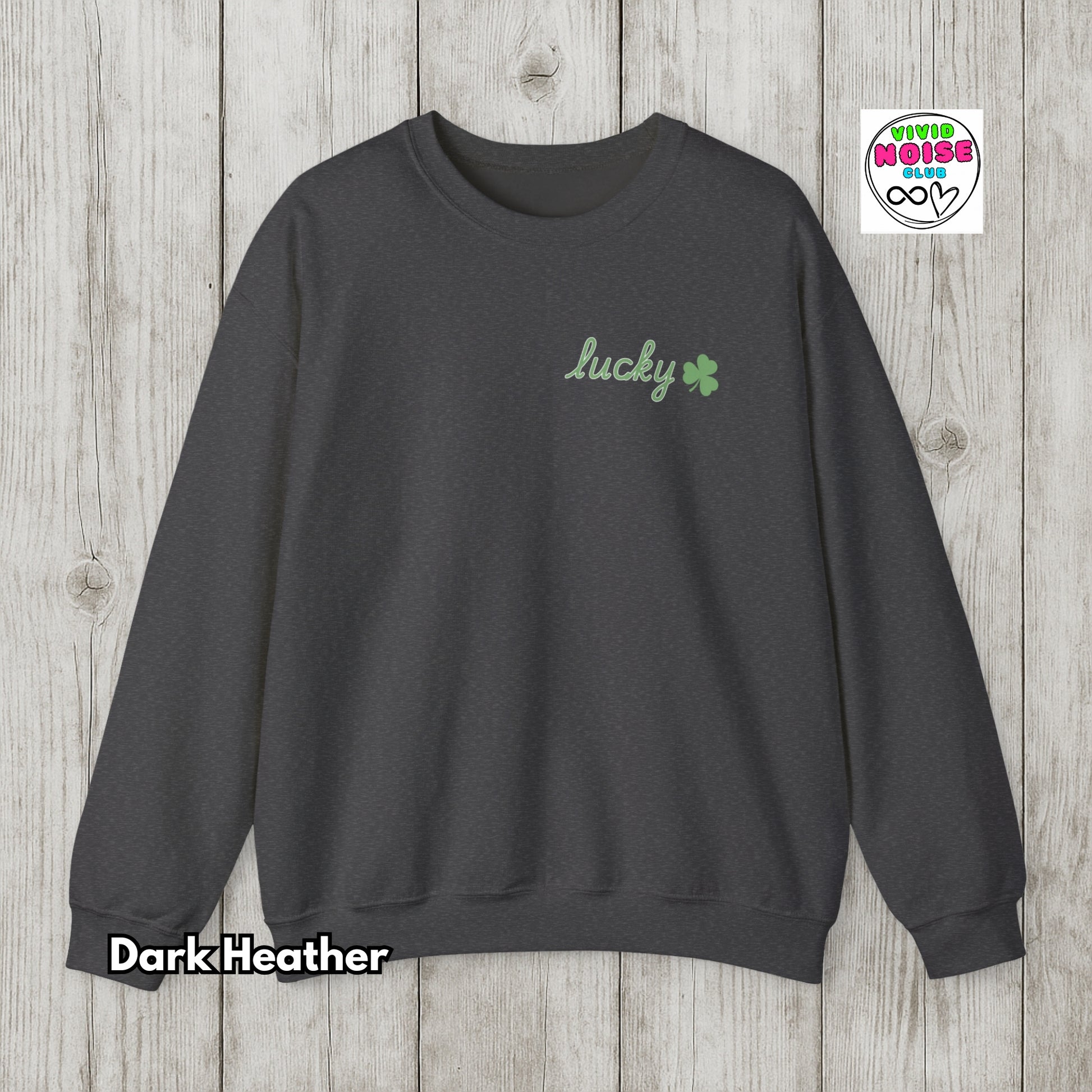 Dark Heather sweatshirt with 'lucky' and clover design on a wooden surface, featuring Vivid Noise logo.