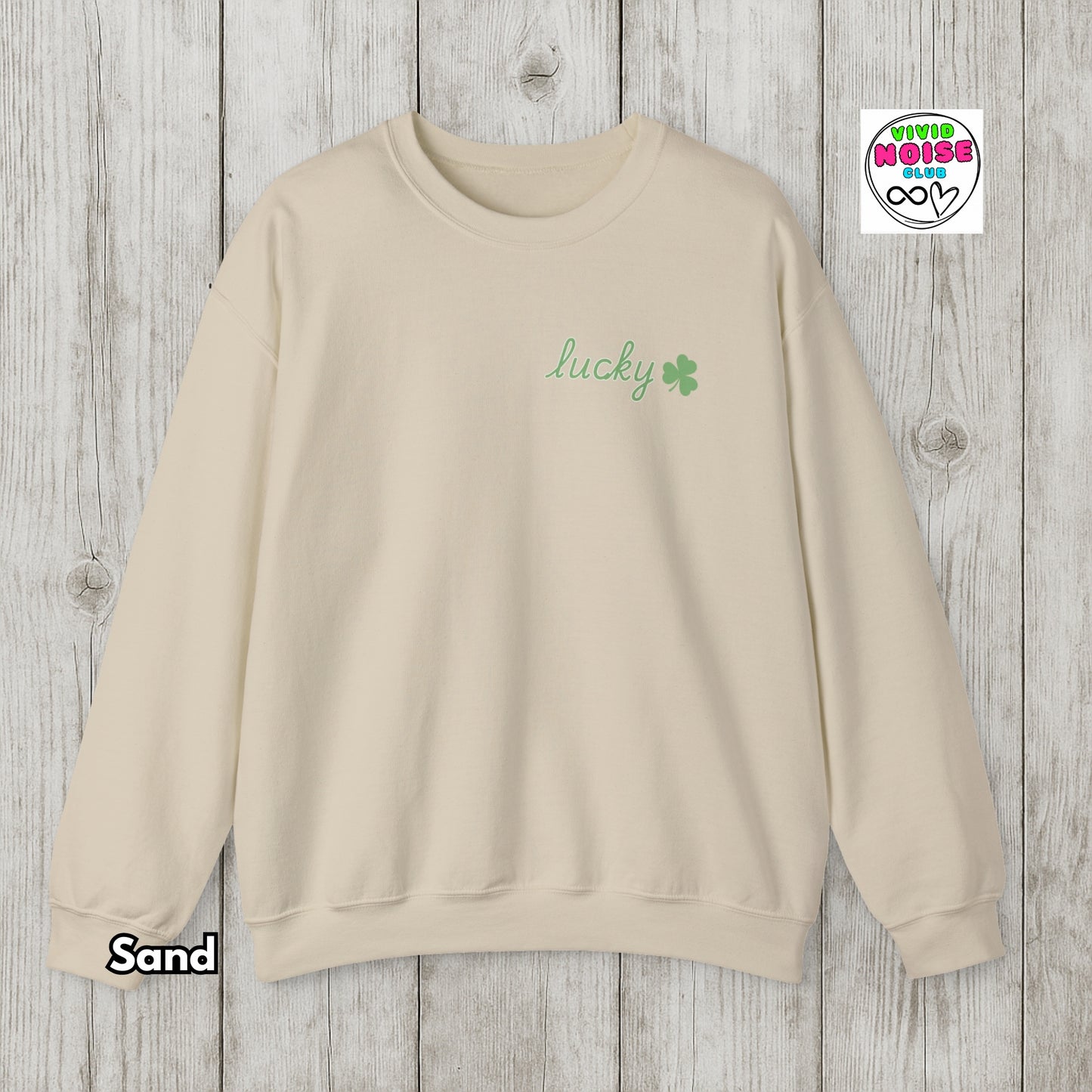 Beige sweatshirt with 'lucky' and clover design on a wooden surface, featuring Vivid Noise logo.