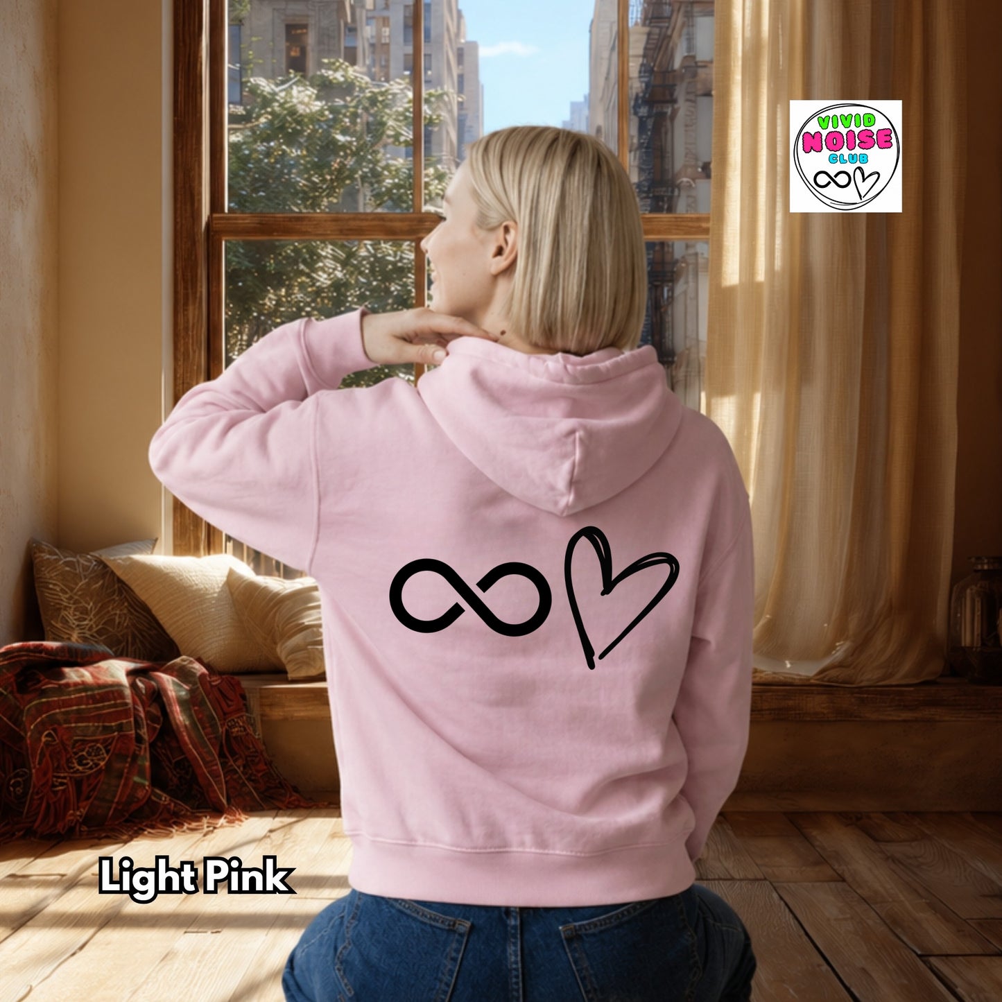Person wearing a light pink hoodie with a heart and infinity symbol on the back, standing in a room with a window and curtains.