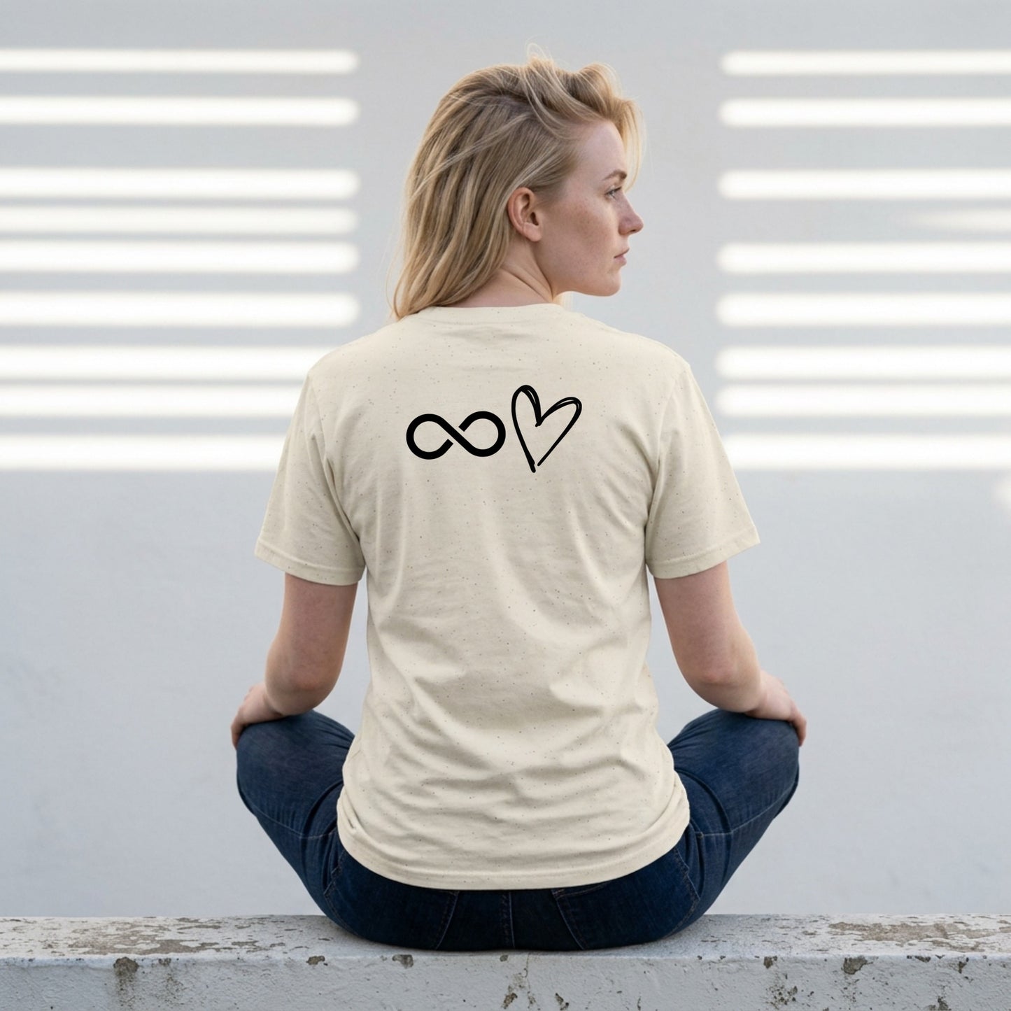 Person wearing a beige t-shirt with a symbol on the back, sitting against a light-colored wall.