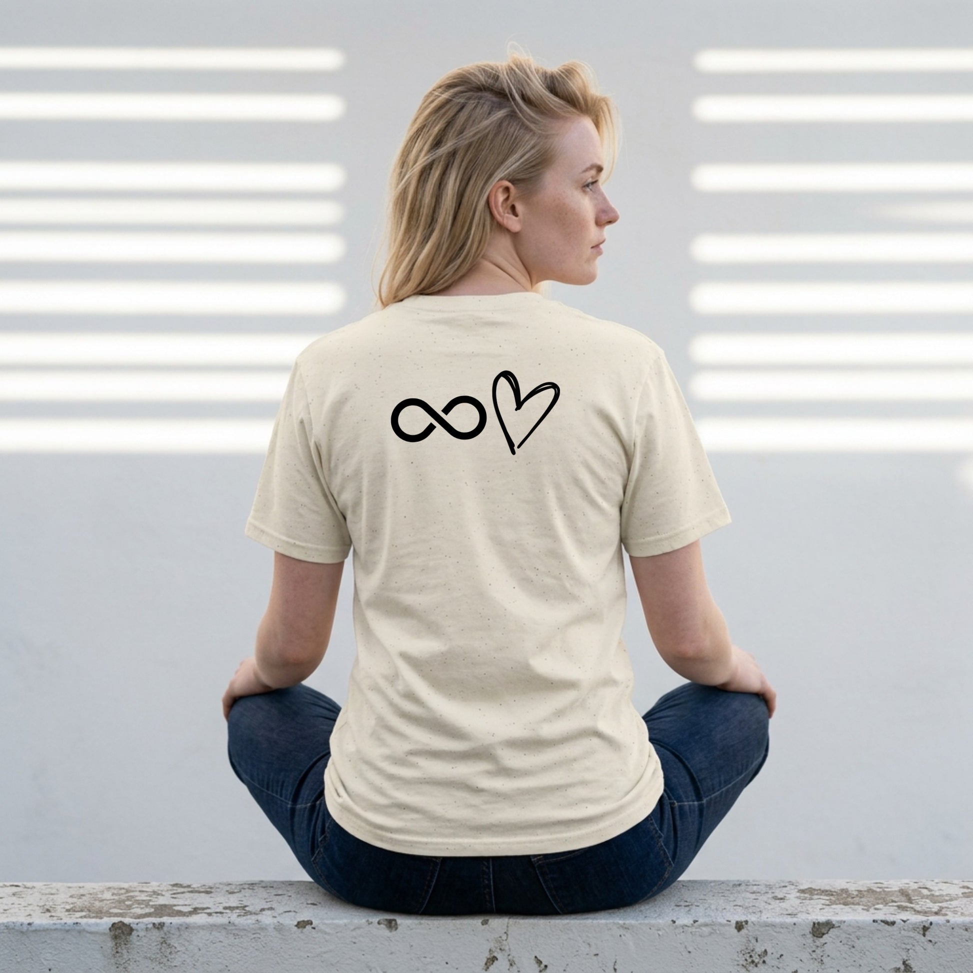 Person wearing a beige t-shirt with a symbol on the back, sitting against a light-colored wall.