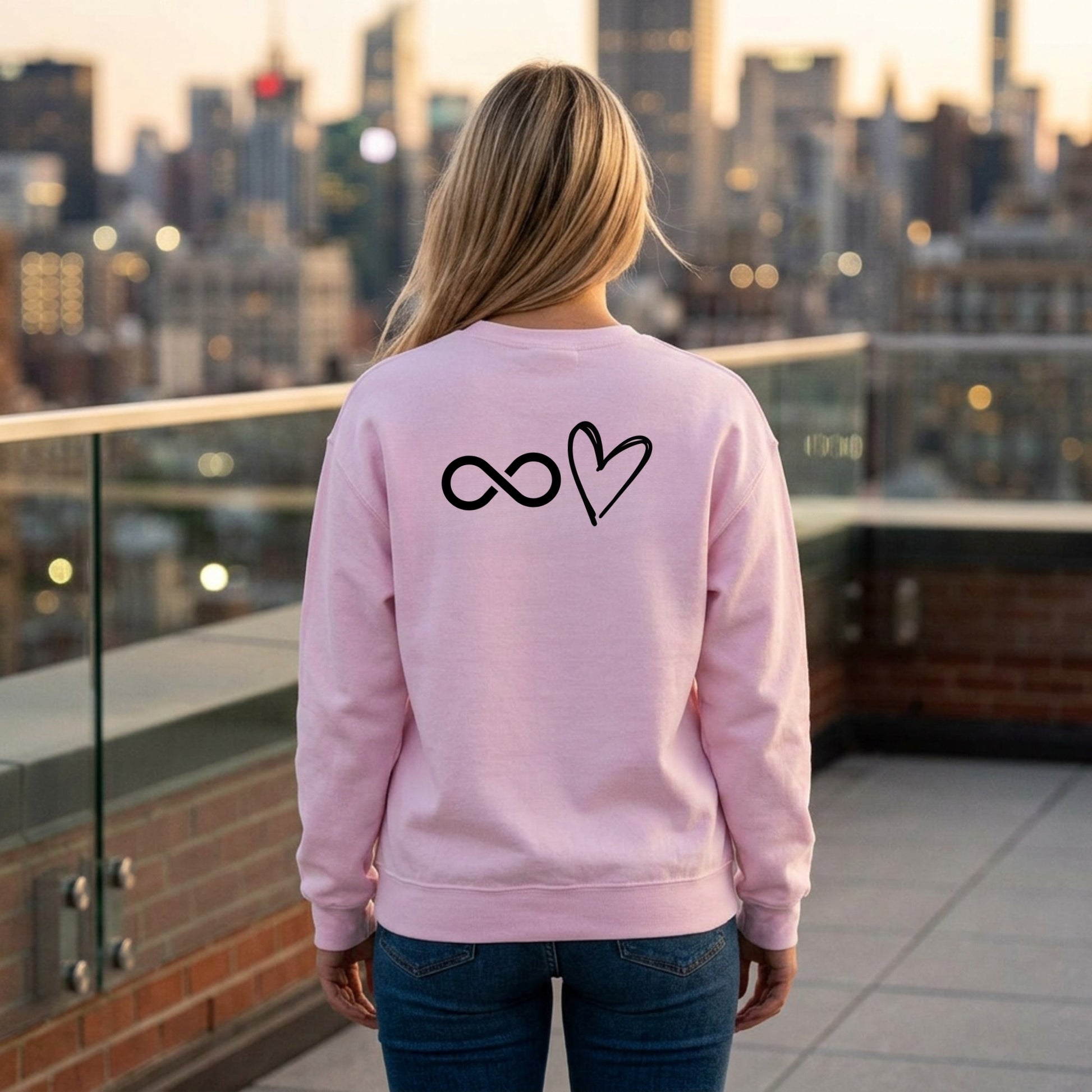 Person wearing a pink sweatshirt with infinity and heart symbols on the back, standing on a rooftop with a cityscape in the background.