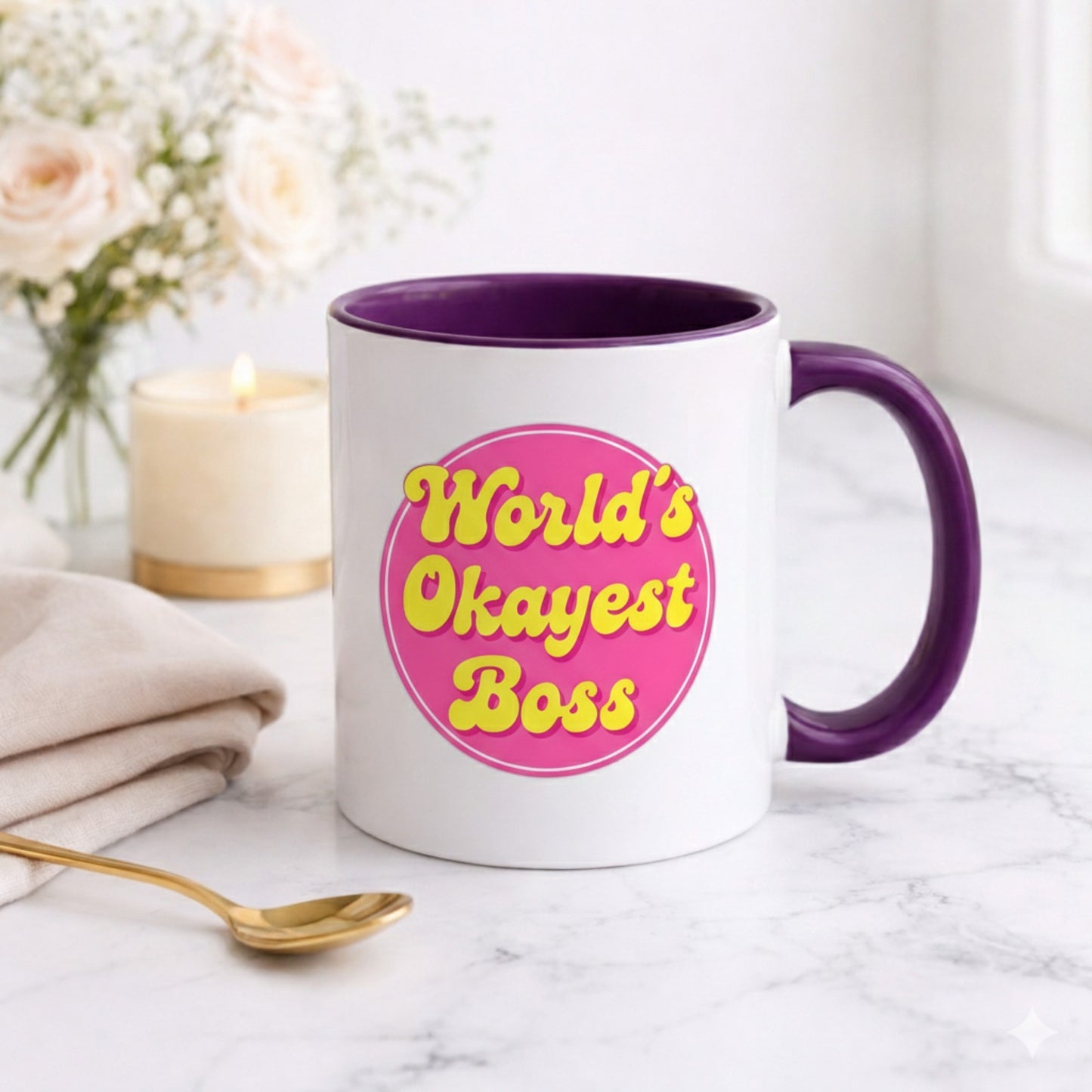 White mug with purple handle and interior, featuring 'World's Okayest Boss' text on a pink background, placed on a marble surface with flowers and a candle in the background.