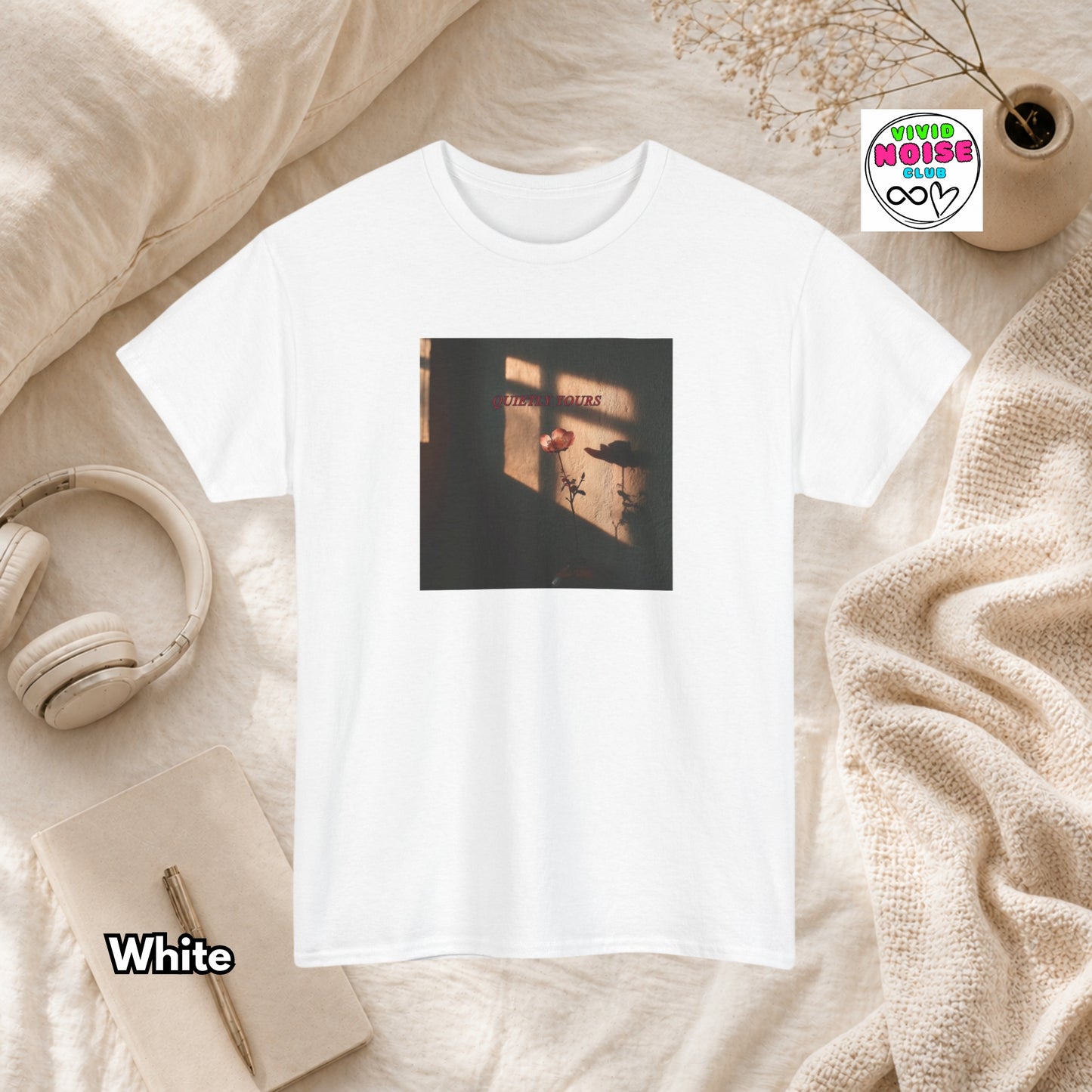 White t-shirt with Quietly Yours floral graphic design on a beige surface with headphones and a notebook.