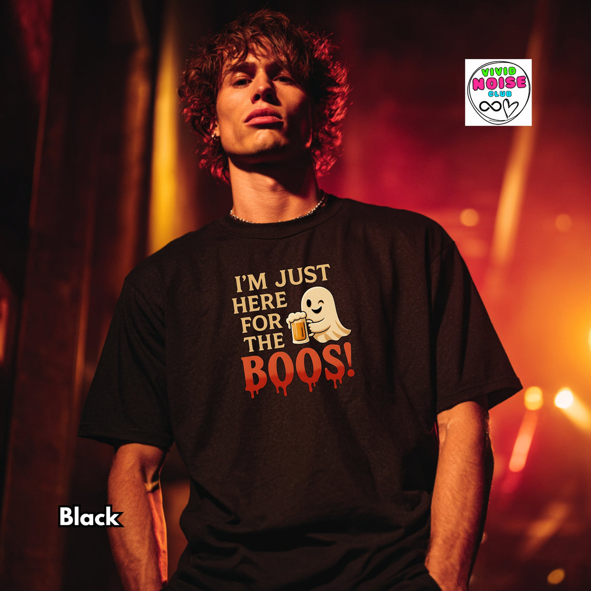 A man wearing a black graphic t-shirt with a ghost and red text that reads 'I'M JUST HERE FOR THE BOOS!'. The background is a warm, orange-toned stage or club setting.