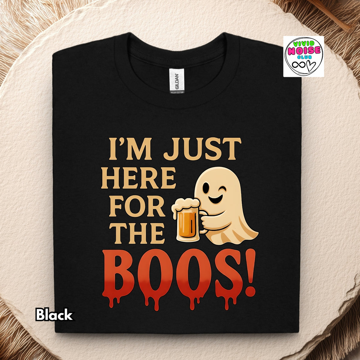 A black short-sleeve t-shirt with a Halloween graphic print reading 'I'M JUST HERE FOR THE BOOS!' in gold and dripping red-orange text, featuring a cartoon ghost holding a beer mug. The shirt is displayed flat on a textured beige wood slice background with a brand logo in the top right corner.