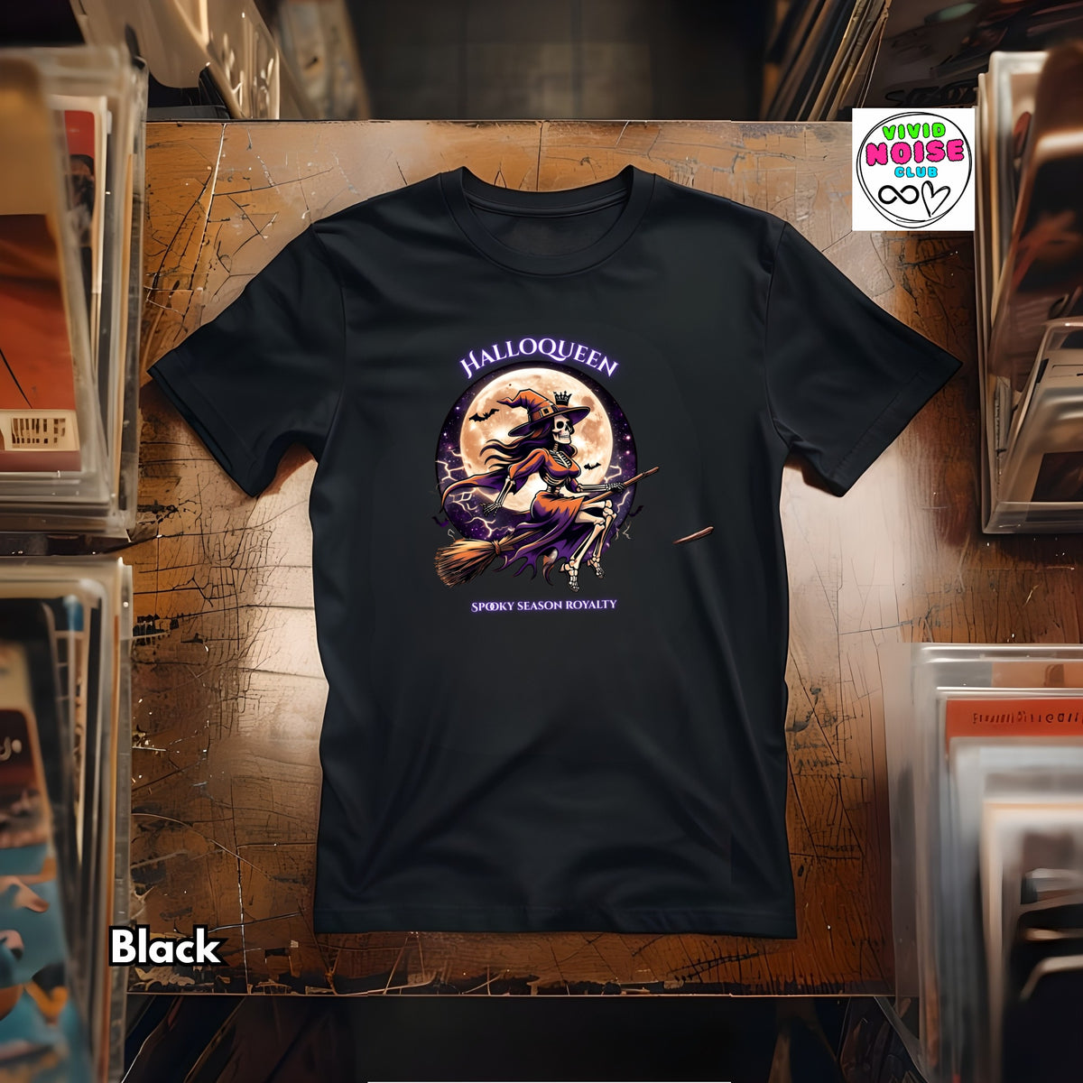 A black short-sleeve graphic T-shirt laid flat on a record store counter surface. The shirt has a Halloween-themed front graphic with the words 'HALLOQUEEN' and 'SPOOKY SEASON ROYALTY' and an illustration of a witch riding a broomstick in front of a full moon.