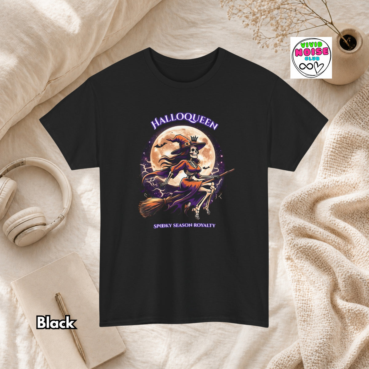 A black short-sleeve graphic T-shirt laid flat on a cream fabric surface with styled props including headphones, a notebook, a candle, and a small plant. The shirt has a Halloween-themed front graphic with the words 'HALLOQUEEN' and 'SPOOKY SEASON ROYALTY' and an illustration of a witch riding a broomstick in front of a full moon.