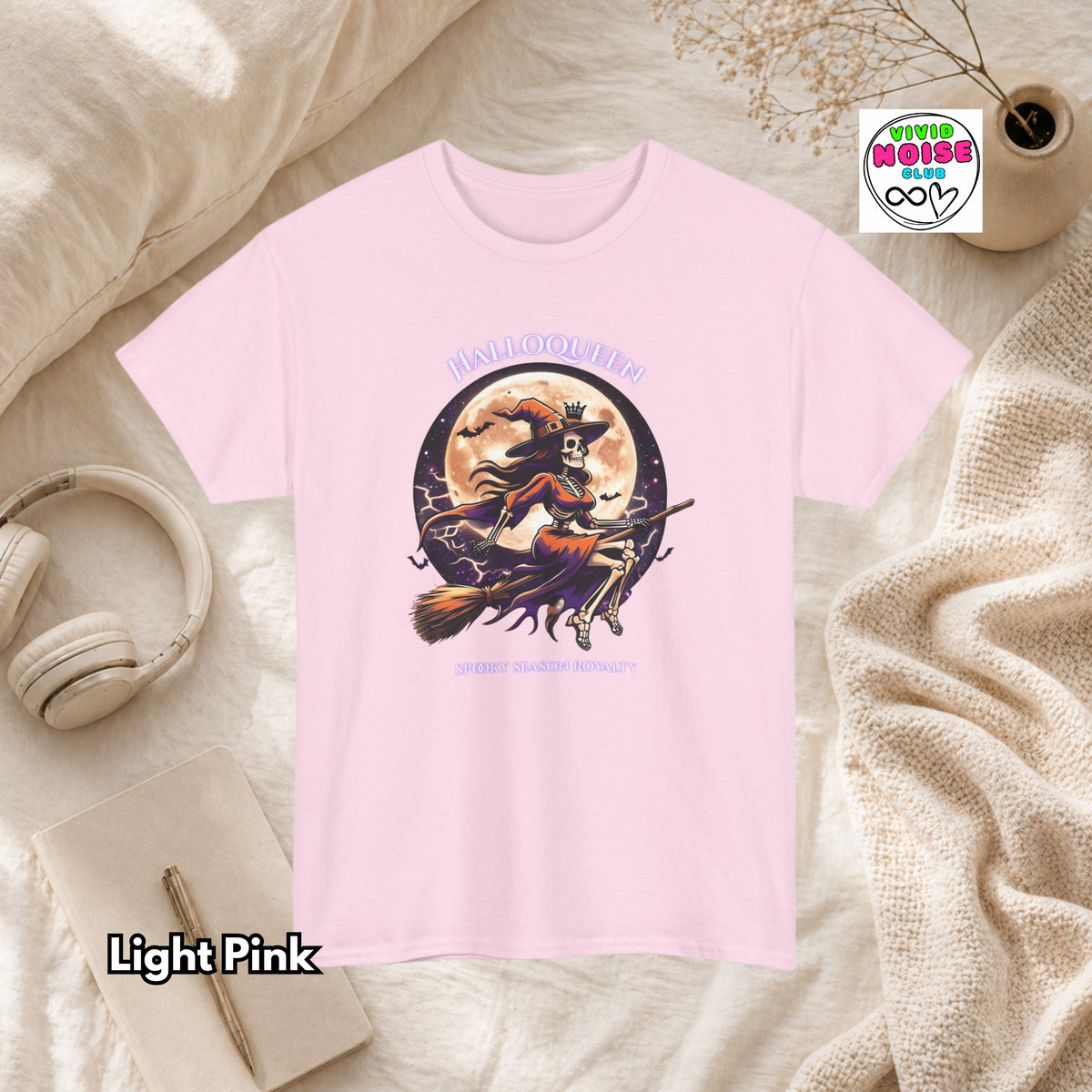 A light pink short-sleeve graphic T-shirt laid flat on a cream fabric surface with styled props including headphones, a notebook, a candle, and a small plant. The shirt has a Halloween-themed front graphic with the words 'HALLOQUEEN' and 'SPOOKY SEASON ROYALTY' and an illustration of a witch riding a broomstick in front of a full moon.