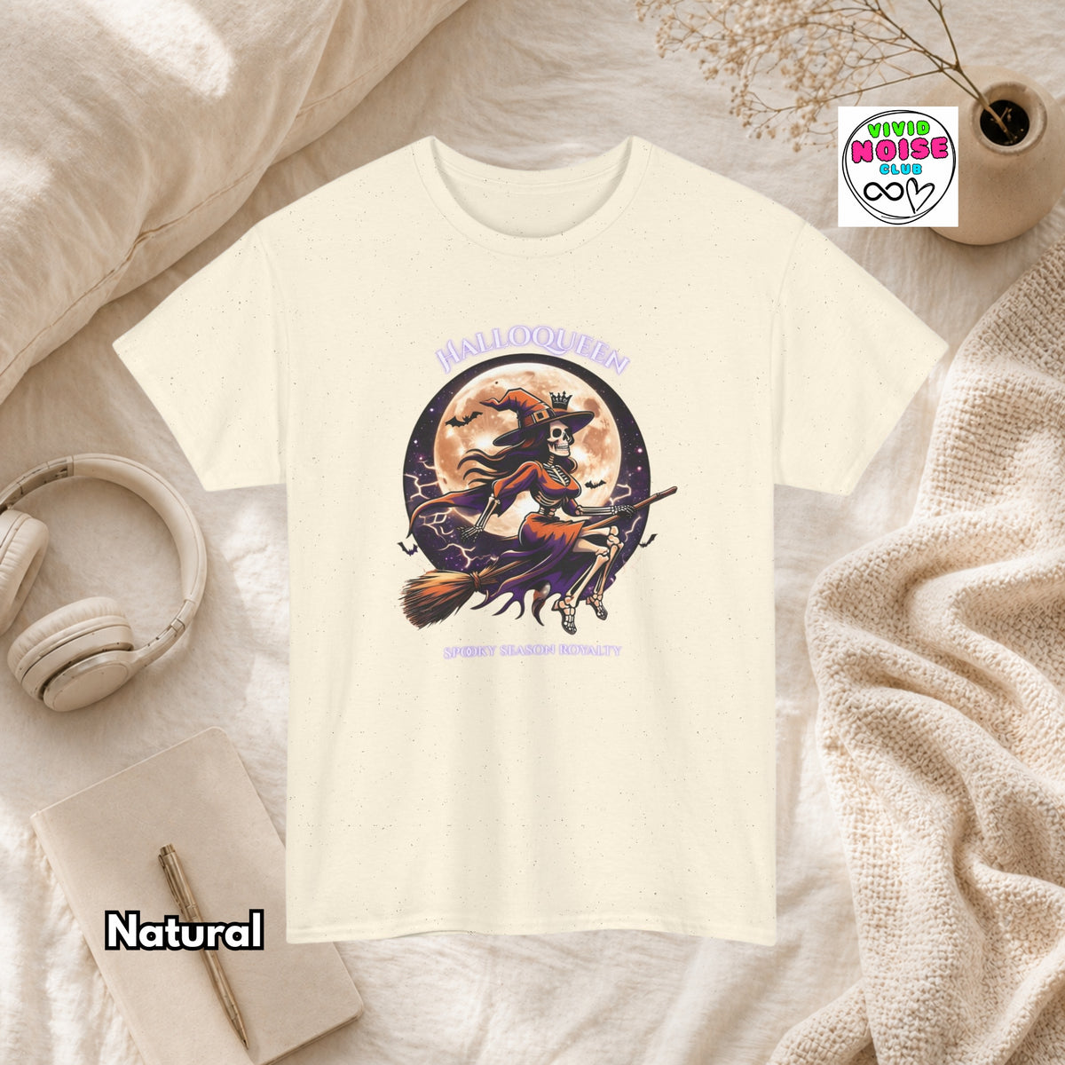 A cream short-sleeve graphic T-shirt laid flat on a cream fabric surface with styled props including headphones, a notebook, a candle, and a small plant. The shirt has a Halloween-themed front graphic with the words 'HALLOQUEEN' and 'SPOOKY SEASON ROYALTY' and an illustration of a witch riding a broomstick in front of a full moon.