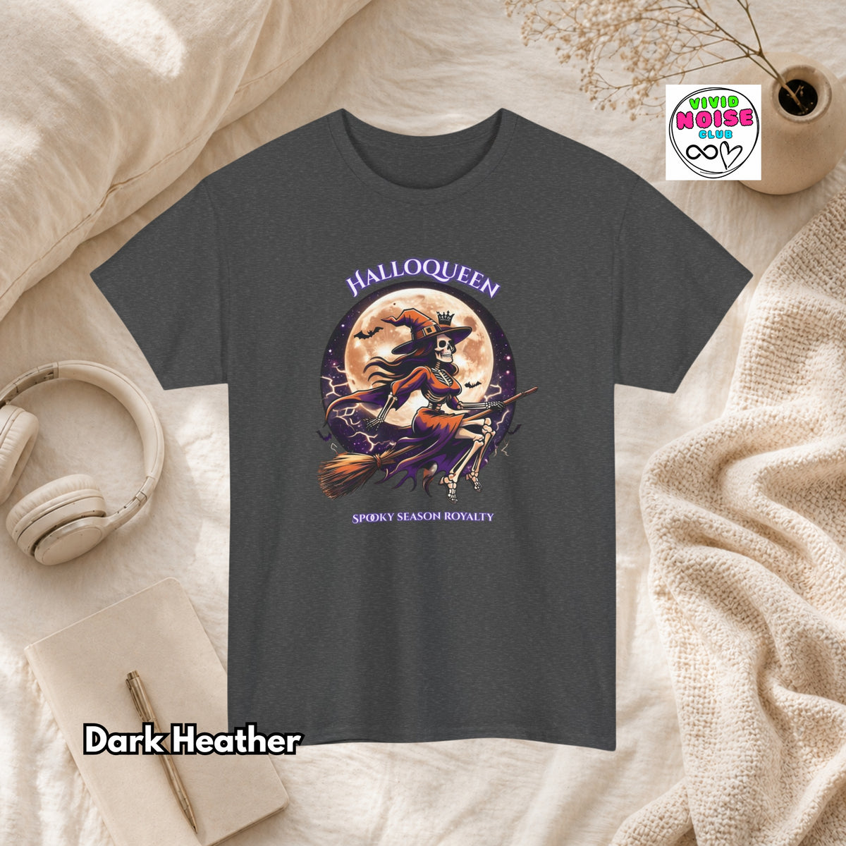 A dark heather gray short-sleeve graphic T-shirt laid flat on a cream fabric surface with styled props including headphones, a notebook, a candle, and a small plant. The shirt has a Halloween-themed front graphic with the words 'HALLOQUEEN' and 'SPOOKY SEASON ROYALTY' and an illustration of a witch riding a broomstick in front of a full moon.