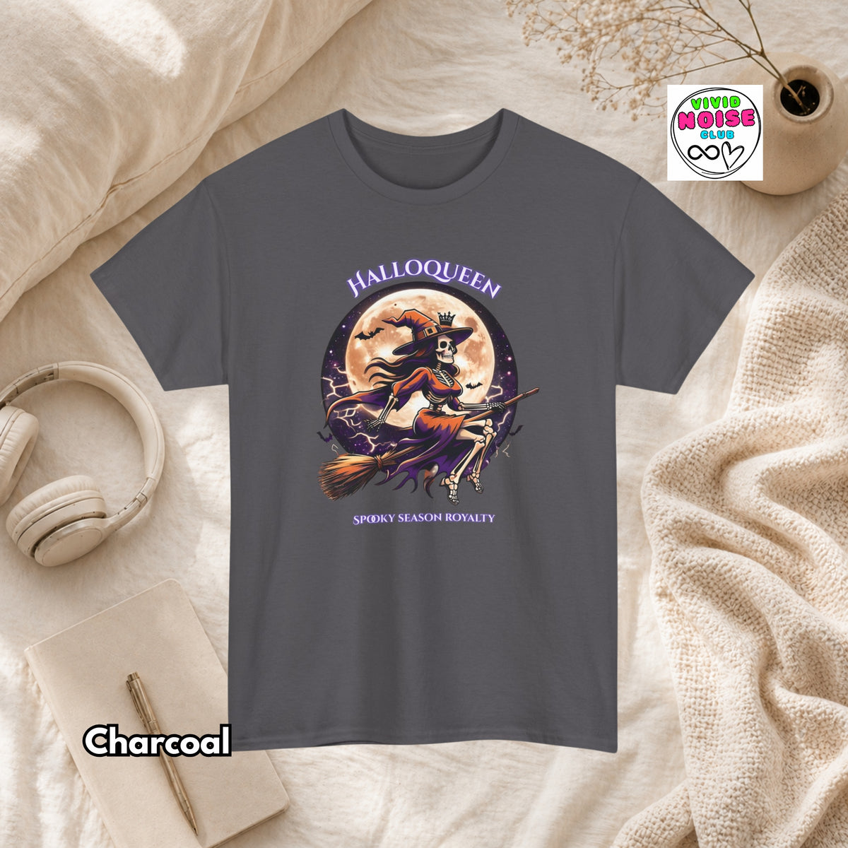 A charcoal gray short-sleeve graphic T-shirt laid flat on a cream fabric surface with styled props including headphones, a notebook, a candle, and a small plant. The shirt has a Halloween-themed front graphic with the words 'HALLOQUEEN' and 'SPOOKY SEASON ROYALTY' and an illustration of a witch riding a broomstick in front of a full moon.