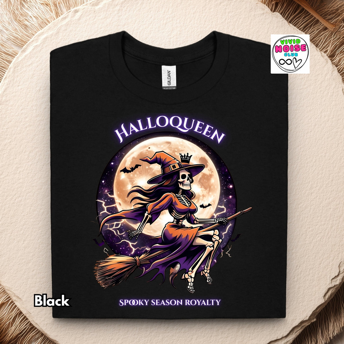 A black short-sleeve t-shirt with a colorful graphic of a skeleton witch riding a broomstick in front of a full moon, with the text 'HALLOOQUEEN' and 'SPOOKY SEASON ROYALTY'. The shirt is displayed flat on a textured beige surface.