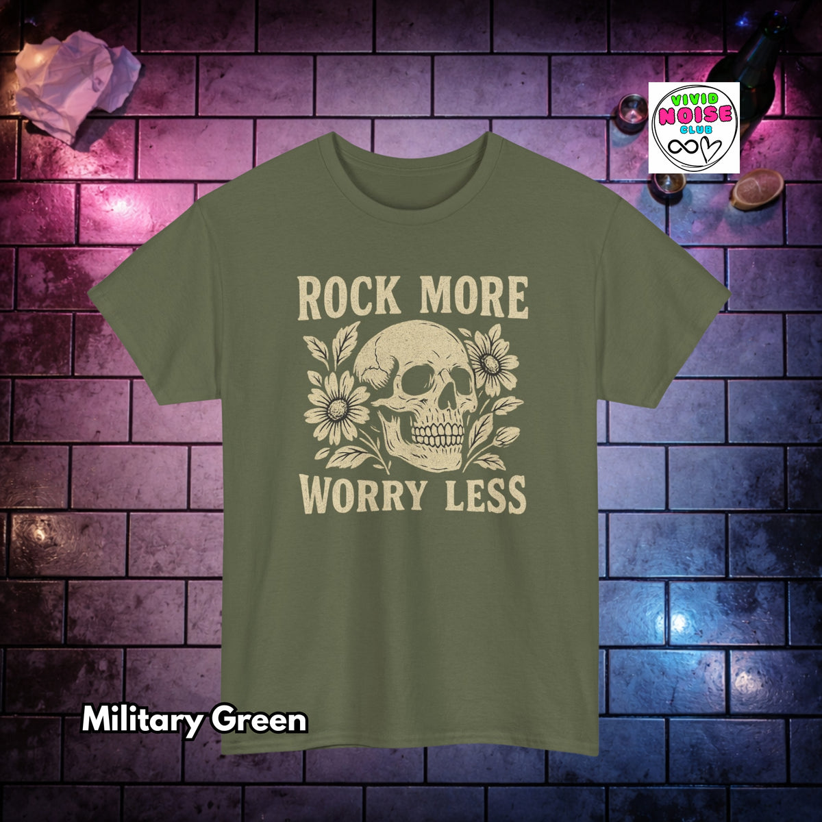 A military green short-sleeve t-shirt with a beige graphic of a skull and flowers and the text 'ROCK MORE WORRY LESS' on the front, displayed against a purple-lit brick wall background.