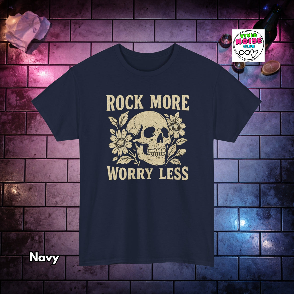 A navy blue short-sleeve graphic t-shirt featuring a cream-colored skull and floral design with the text 'ROCK MORE WORRY LESS'. The shirt is displayed against a dark brick wall background with purple and blue lighting, with a 'Vivid Noise Club' brand logo in the top right and a 'Navy' color label in the bottom left.
