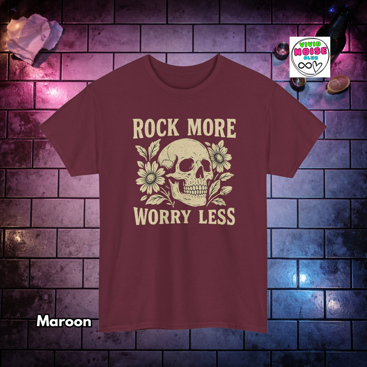 A maroon short-sleeve t-shirt with a cream graphic of a skull and flowers and the text 'ROCK MORE WORRY LESS', displayed against a dark brick wall background with purple and blue lighting.