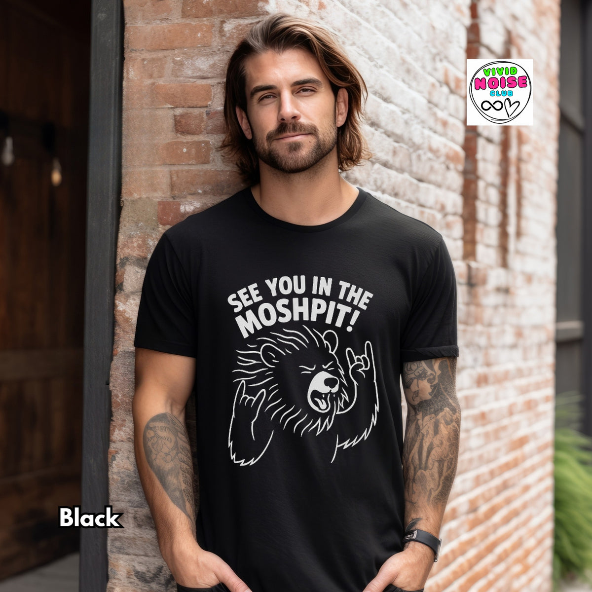 A man with tattoos stands against a brick wall wearing a black graphic t-shirt with a white bear illustration and the text 'SEE YOU IN THE MOSHPIT!'. A 'White Moose Co' logo is visible in the top right, and 'Black' is written in the bottom left.