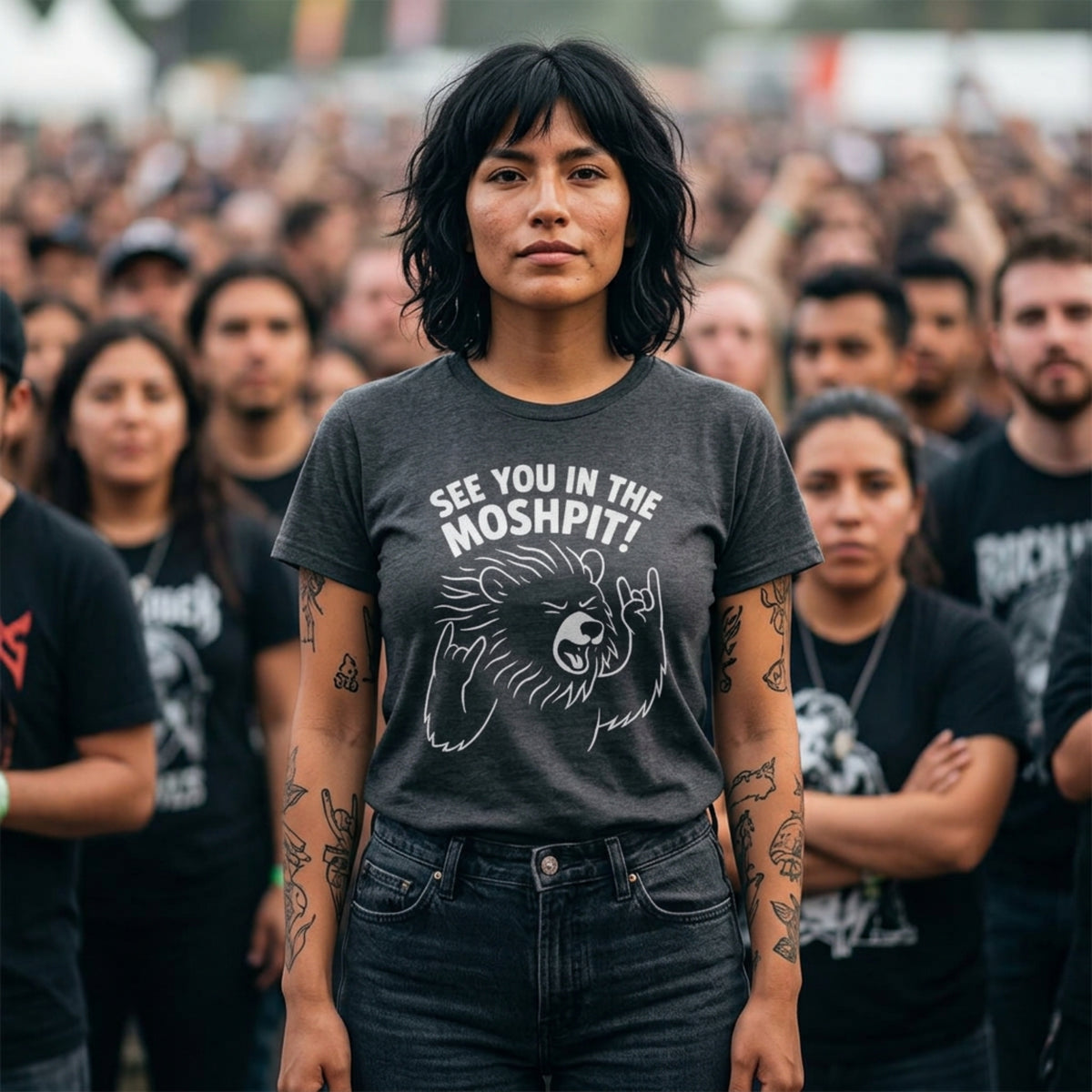 A woman standing in a crowd wearing a dark gray graphic t-shirt with the text 'SEE YOU IN THE MOSHPIT!' and a bear illustration, paired with dark wash jeans.