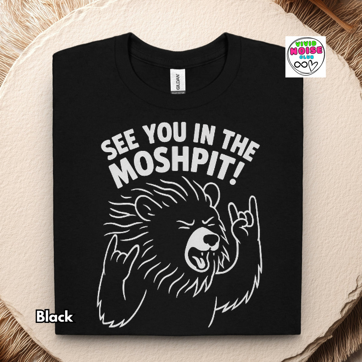 A folded black t-shirt with a white graphic of a bear making a 'I love you' hand gesture and the text 'SEE YOU IN THE MOSHPIT!'. The shirt is displayed flat on a textured beige stone slab with a faux fur background.