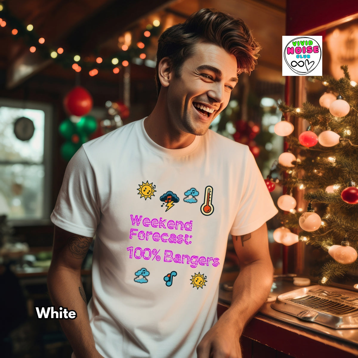 A man wearing a white graphic t-shirt with a colorful 'Weekend Forecast: 100% Bangers' print, standing in a cozy, festive room with a Christmas tree and warm string lights. A 'White' color label is in the bottom-left corner, and a 'Little Miss Cozy' brand logo is in the top-right.