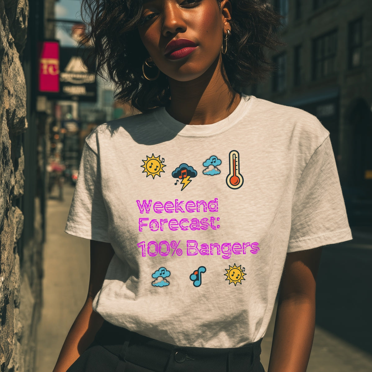 A woman wearing a light gray heather t-shirt with a colorful graphic that reads 'Weekend Forecast: 100% Bangers' with weather icons, standing in an outdoor urban setting.
