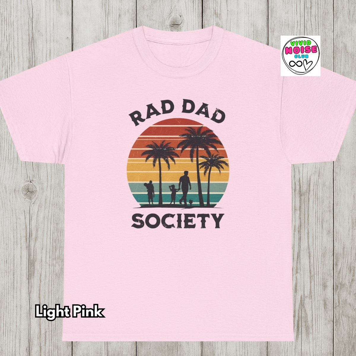A light pink short-sleeve t-shirt laid flat on a light gray wood plank background. The shirt features a retro-style graphic with the text 'RAD DAD SOCIETY' surrounding a sunset illustration with palm trees and silhouettes of people.