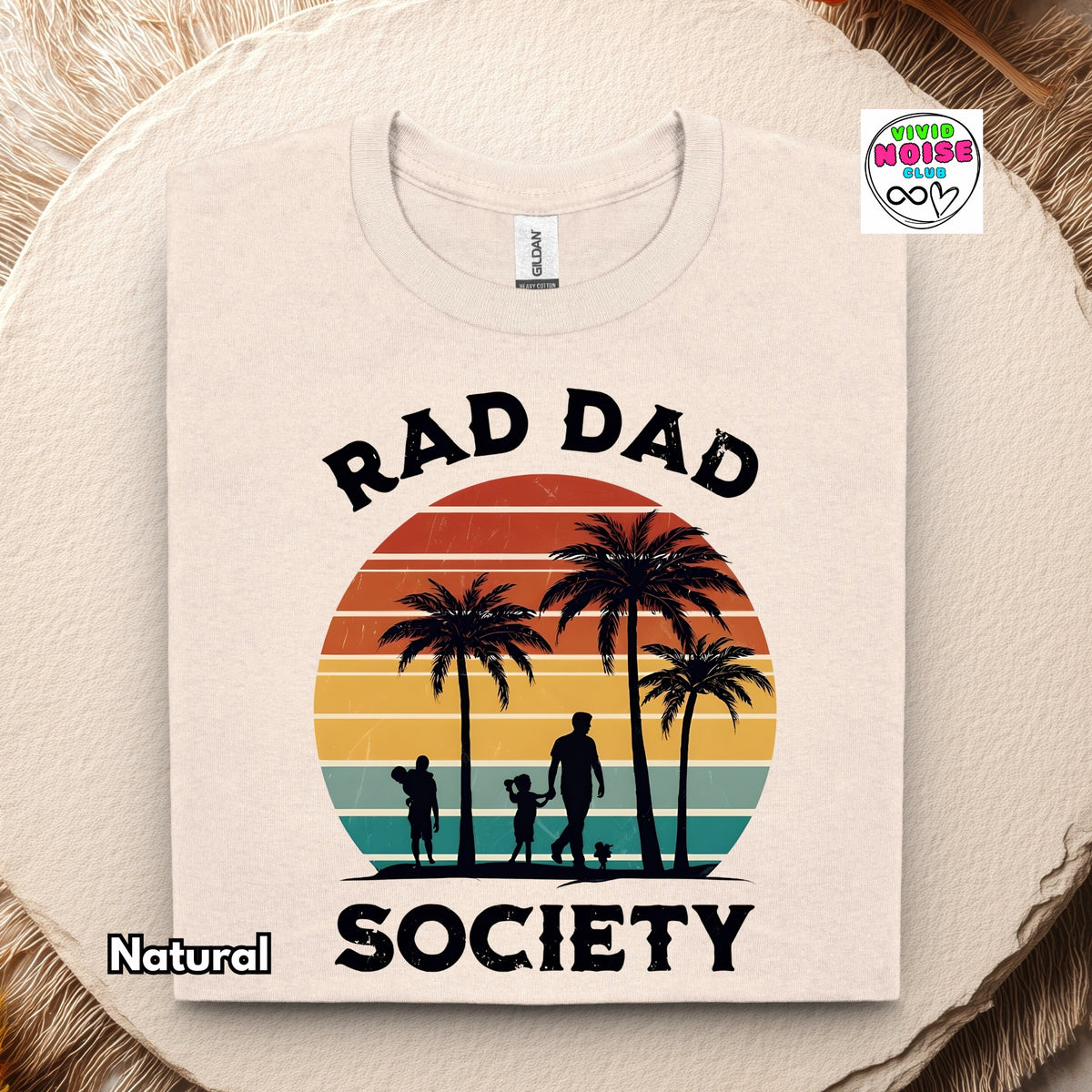 A beige crewneck t-shirt with a retro sunset graphic and the text 'RAD DAD SOCIETY' printed on the front, displayed on a rustic wood slice background.