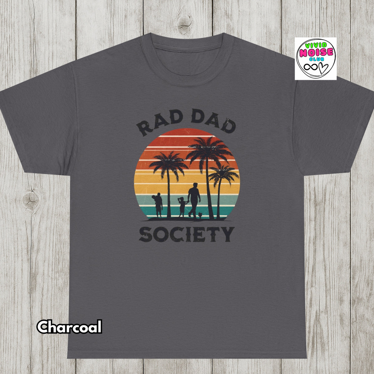 A charcoal gray short-sleeve t-shirt with a retro sunset graphic featuring palm trees and silhouettes of a man and dog, with the text 'RAD DAD SOCIETY'. The shirt is displayed flat on a light wood background, with a 'Charcoal' color label in the bottom-left corner and a 'Vivid Noise Club' logo in the top right corner.