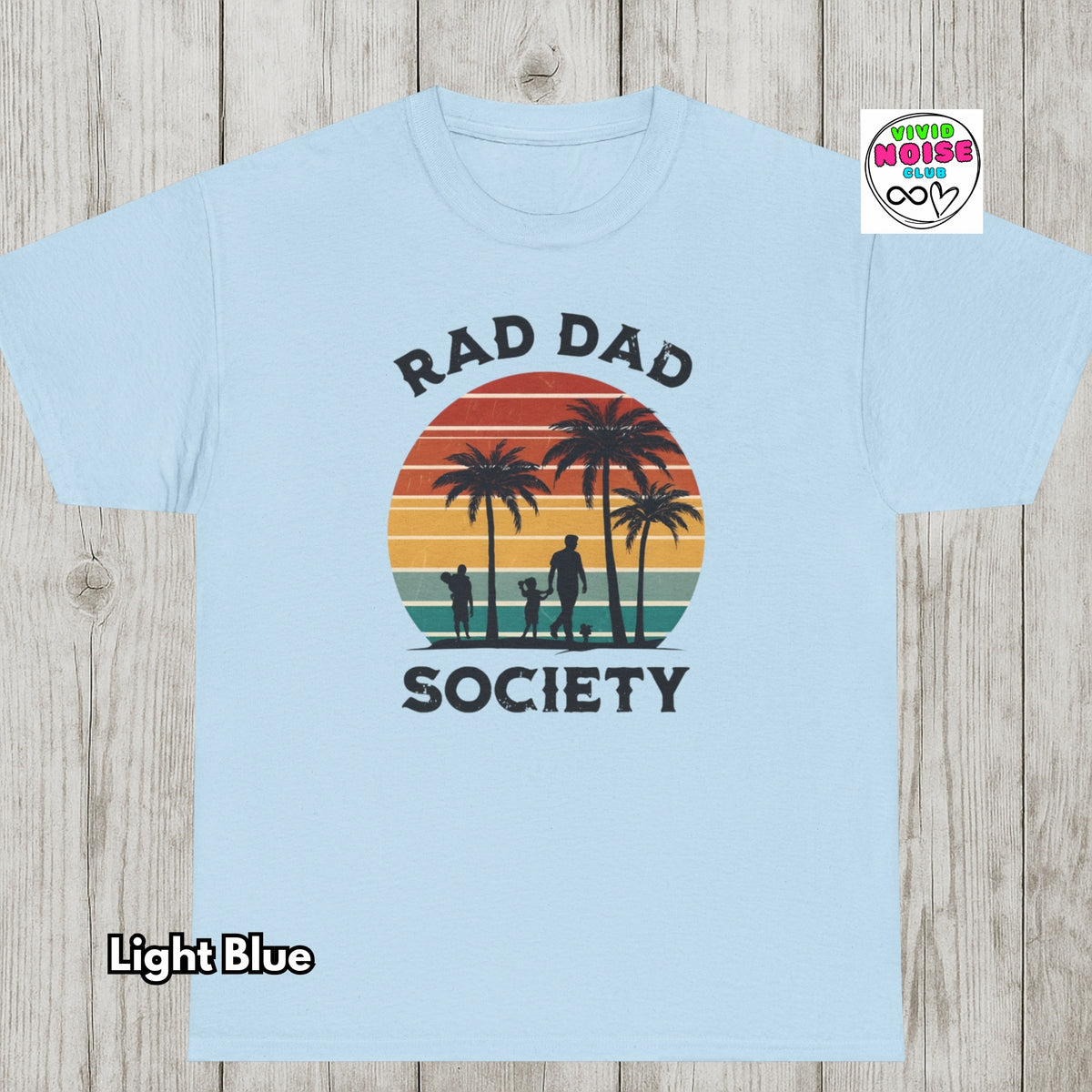 A light blue crewneck t-shirt with a retro sunset graphic and the text 'RAD DAD SOCIETY' printed on the front, displayed flat on a light wood background with a 'Vivid Noise Club' badge in the top right corner.