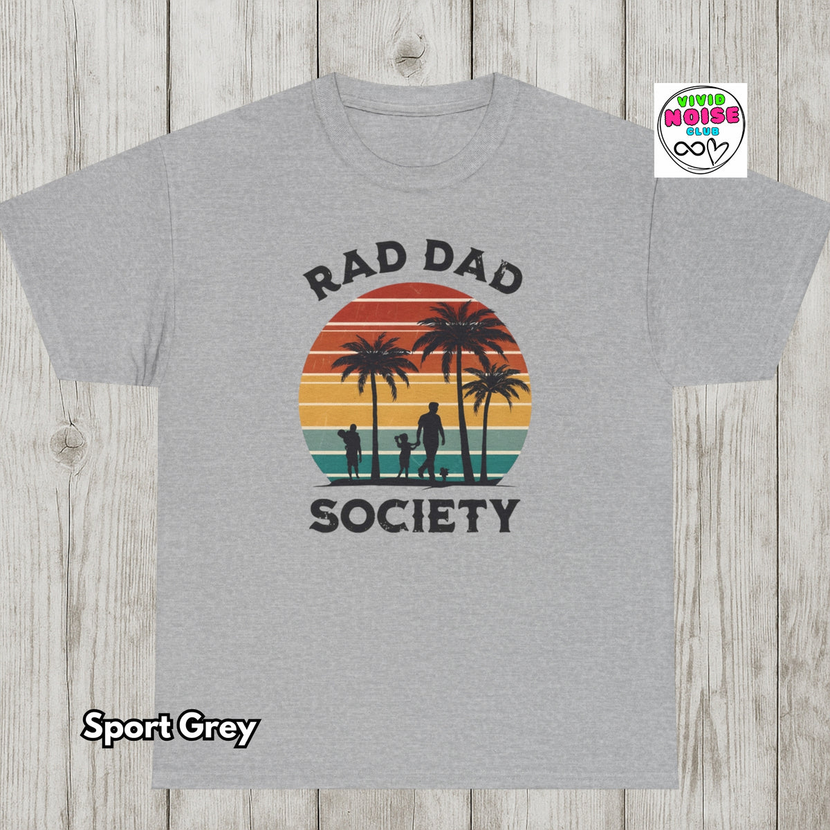 A sport grey t-shirt with a retro sunset graphic featuring palm trees and silhouettes of a dad and dog, with the text 'RAD DAD SOCIETY'. The t-shirt is displayed flat on a light grey wood plank background, with a 'Vivid Noise Club' logo in the top right and 'Sport Grey' text in the bottom left.