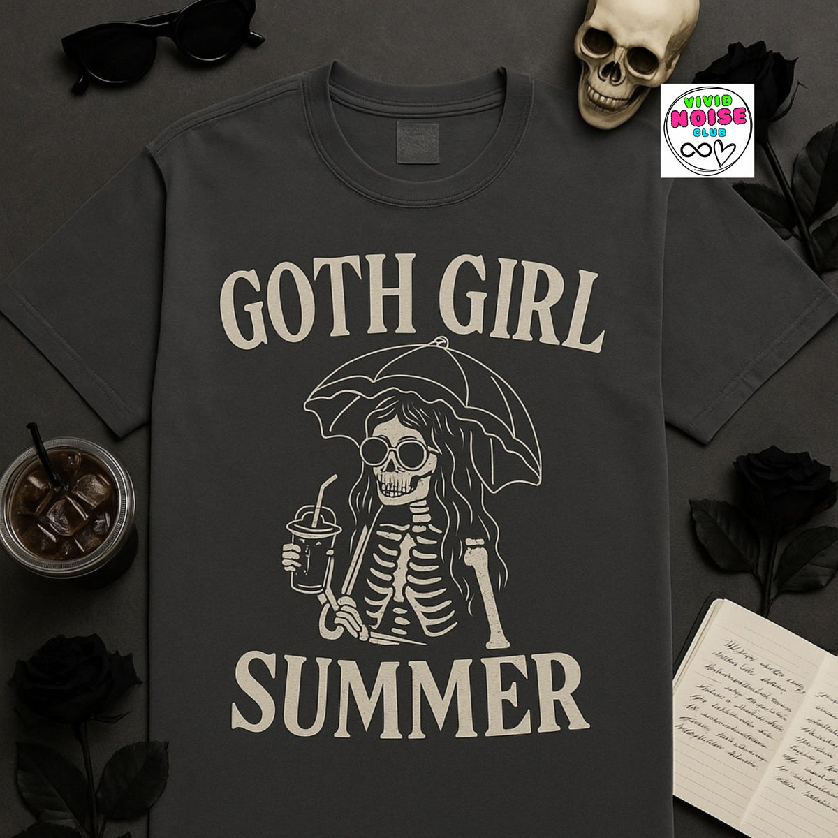 A dark charcoal gray t-shirt with a cream-colored graphic of a skeleton holding a drink under an umbrella, with the text 'GOTH GIRL SUMMER'. The shirt is presented in a styled flat-lay with black roses, sunglasses, a skull, a notebook, and an iced coffee on a dark gray background.