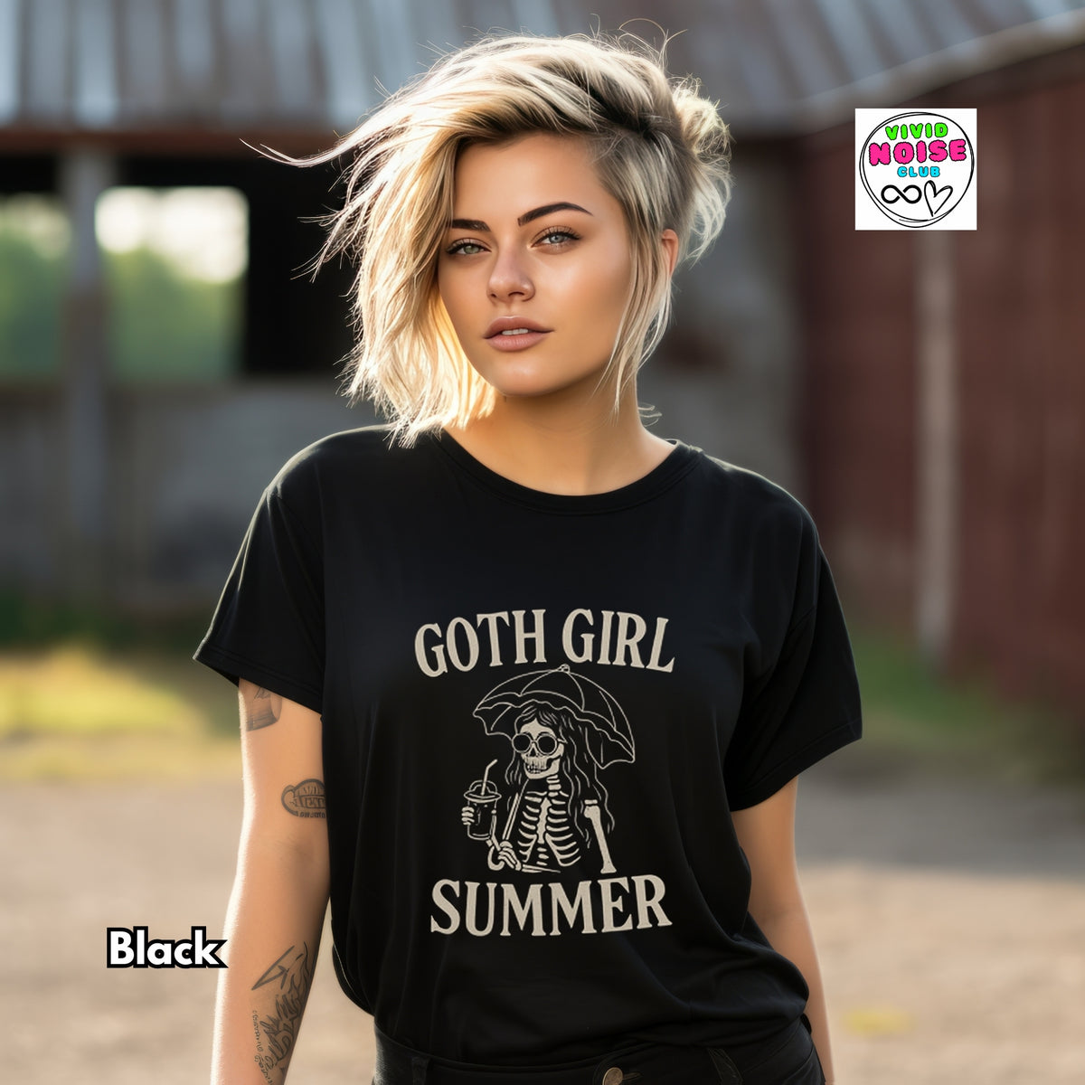 A woman wearing a black graphic t-shirt with a cream-colored gothic skeleton design and the text 'GOTH GIRL SUMMER'. The background is a blurred outdoor setting with rustic barns.