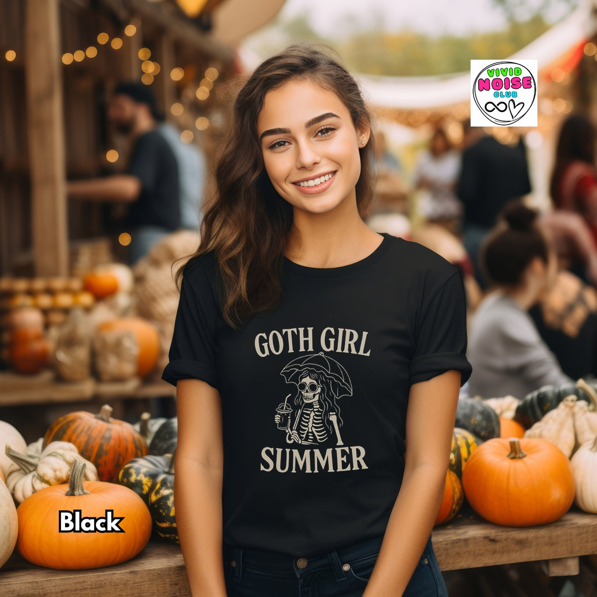 A smiling young woman stands in a fall market setting wearing a black graphic T-shirt with a skeleton holding an umbrella and the text 'GOTH GIRL SUMMER.' Pumpkins and blurred people appear in the background.