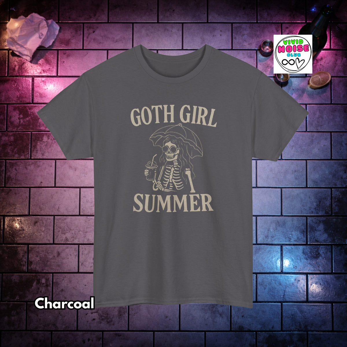 A charcoal grey short-sleeve t-shirt with a cream-colored graphic of a skeleton under an umbrella holding a drink, with the text 'GOTH GIRL SUMMER'. The shirt is displayed against a dark brick wall background with purple and blue lighting, with a 'Vivid Noise Club' logo in the top right and 'Charcoal' text in the bottom left.
