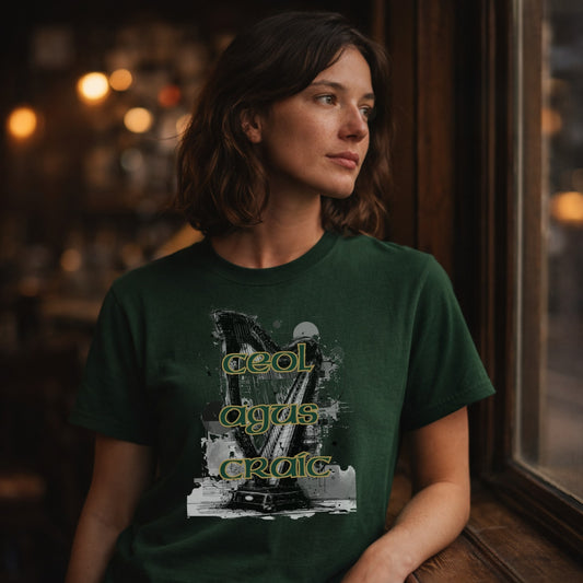 A woman wearing a dark green graphic t-shirt with a stylized graphic of a harp and the text 'Ceol Agus Craic' in yellow-green, posed in a dimly lit, warm-toned indoor setting.
