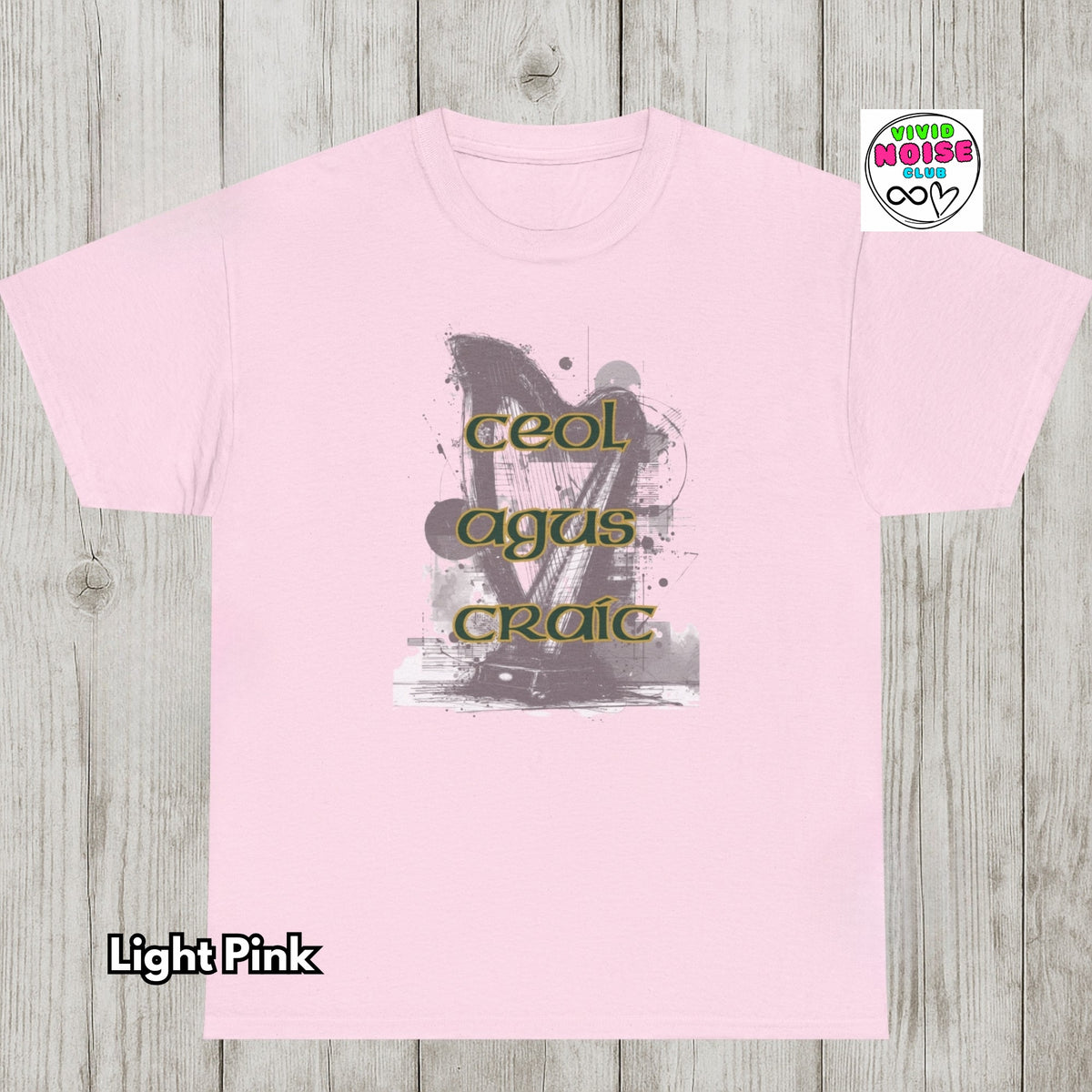 A light pink crewneck t-shirt laid flat on a light gray wood plank background. The center of the shirt features a graphic print with a gray and black abstract design and the words 'ceol agus craic' in olive green.
