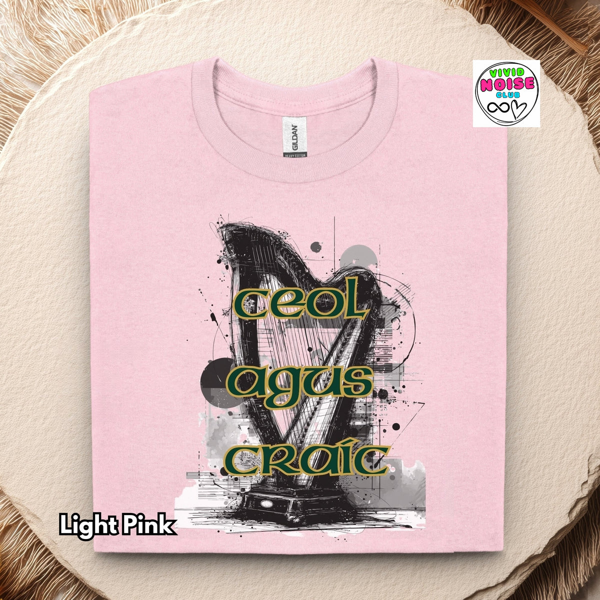 A folded light pink t-shirt with a graphic print of a harp and the Irish Gaelic phrase 'ceol agus craic' in green and gold, displayed on a rustic wood slice background.