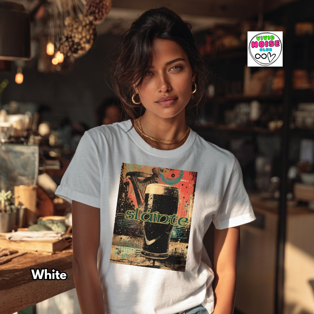 A woman wearing a white graphic t-shirt with a colorful print of a beer glass and the word 'Slainte', set in a warm, dimly lit café environment. A 'White' label is visible in the bottom left, and a 'Vivid Noise Club' logo is in the top right.