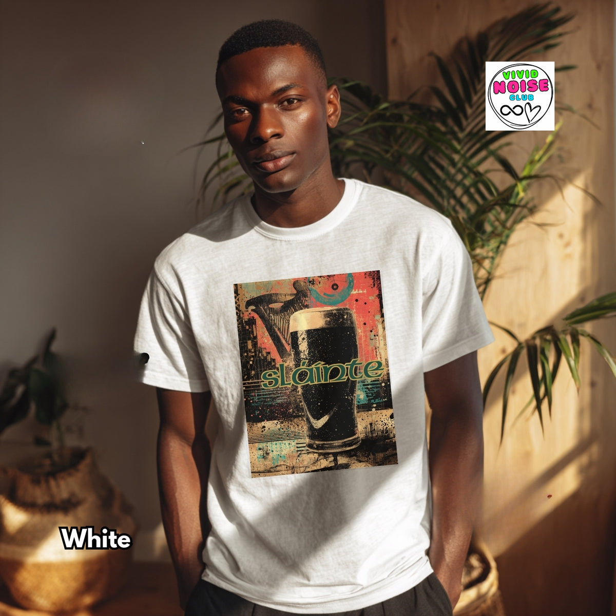 A man wearing a white crewneck t-shirt with a colorful graphic print of a beer mug and the word 'slainte'. The image has a warm, stylized indoor background with plants and a logo overlay in the top right corner.
