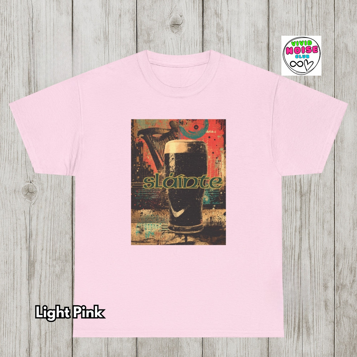 A light pink crewneck t-shirt with a central graphic print of a pint of stout beer and the word 'Slainte' in green, displayed on a gray wood plank background. A 'Vivid Noise Club' logo is in the top right, and 'Light Pink' is written in the bottom left.