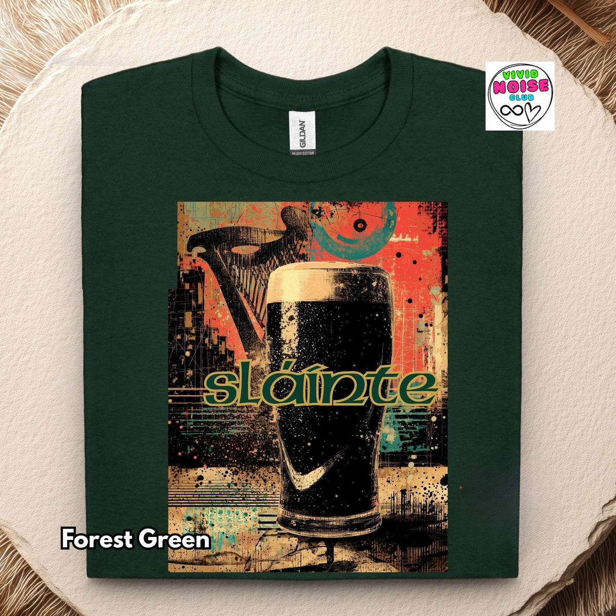 A folded forest green t-shirt with a large graphic print of a pint of black stout and the word 'Sláinte' in green, displayed on a textured beige background with a 'Vivid Noise Club' logo in the top right.