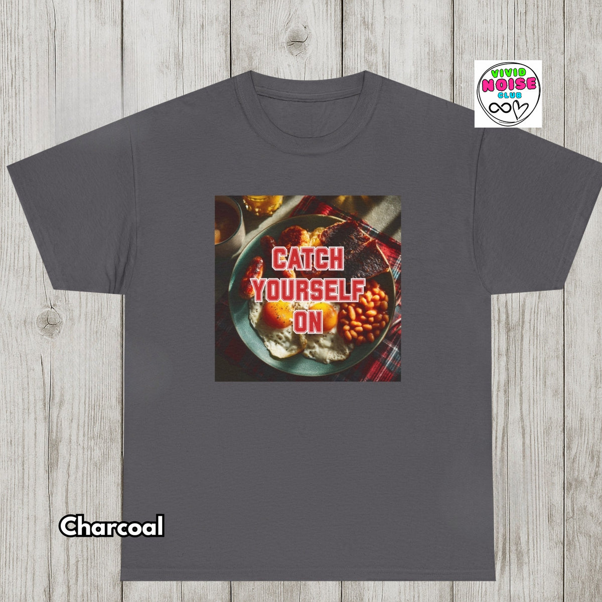 A charcoal gray t-shirt displayed flat against a light wood plank background. The shirt features a square graphic print in the center showing a breakfast plate with eggs, bacon, and beans, overlaid with the text 'CATCH YOURSELF ON' in red.