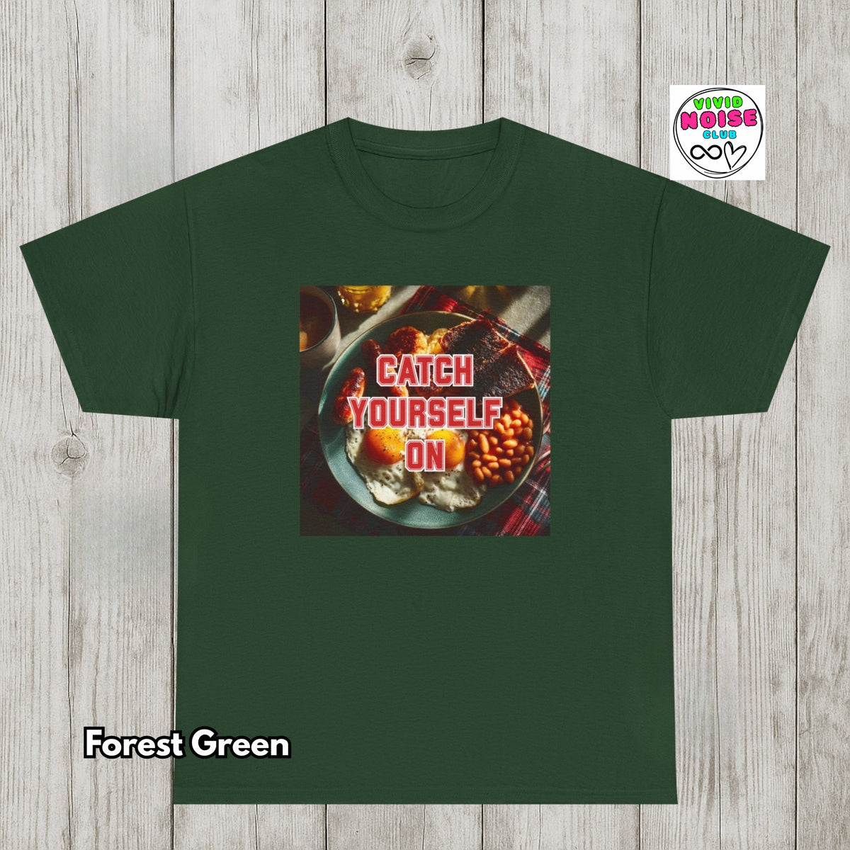 A forest green short-sleeve t-shirt displayed flat against a light gray wood plank background. The center of the shirt features a square graphic print of a breakfast scene with the text 'CATCH YOURSELF ON' in red.