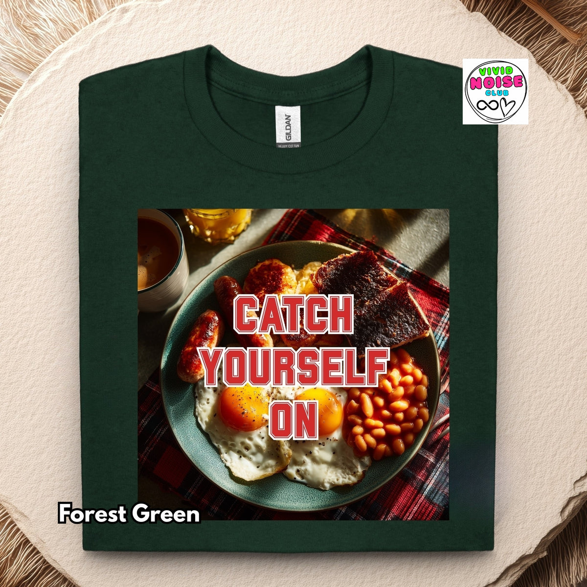A folded forest green t-shirt displayed flat on a textured beige surface. The shirt features a large graphic print of a full English breakfast on a plate with the text 'CATCH YOURSELF ON' in bold red letters.