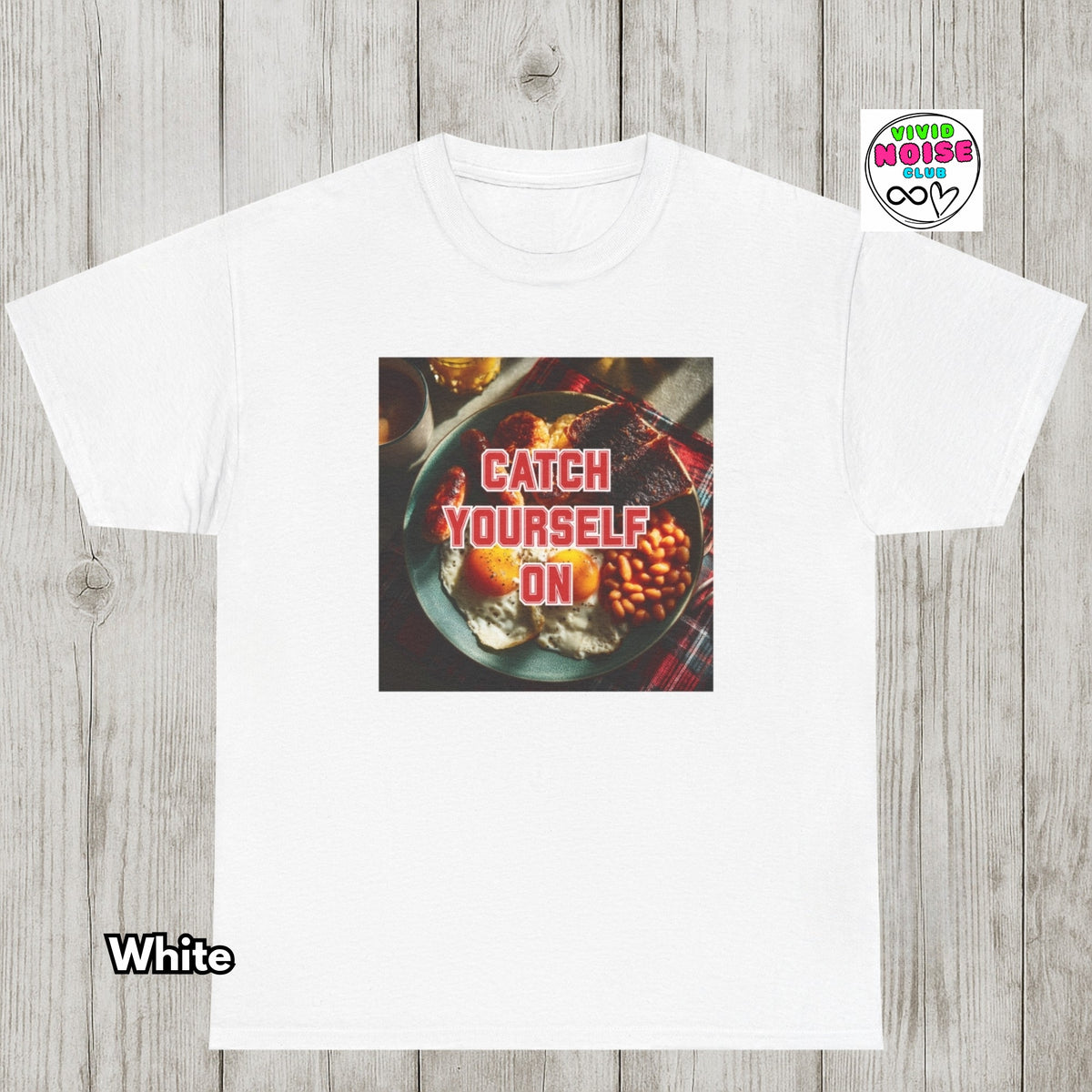 A white short-sleeve t-shirt laid flat on a light gray wood plank background. The center of the shirt features a square graphic print of a plate of food with the text 'CATCH YOURSELF ON' in red.