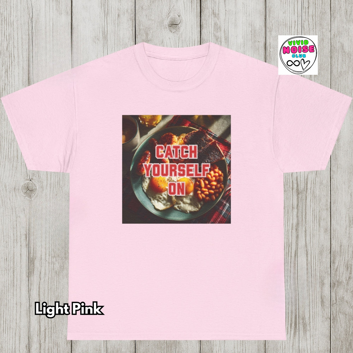 A light pink short-sleeve t-shirt laid flat on a light gray wood plank background. The center of the shirt features a square graphic print of a plate of food with the text 'CATCH YOURSELF ON' in red.