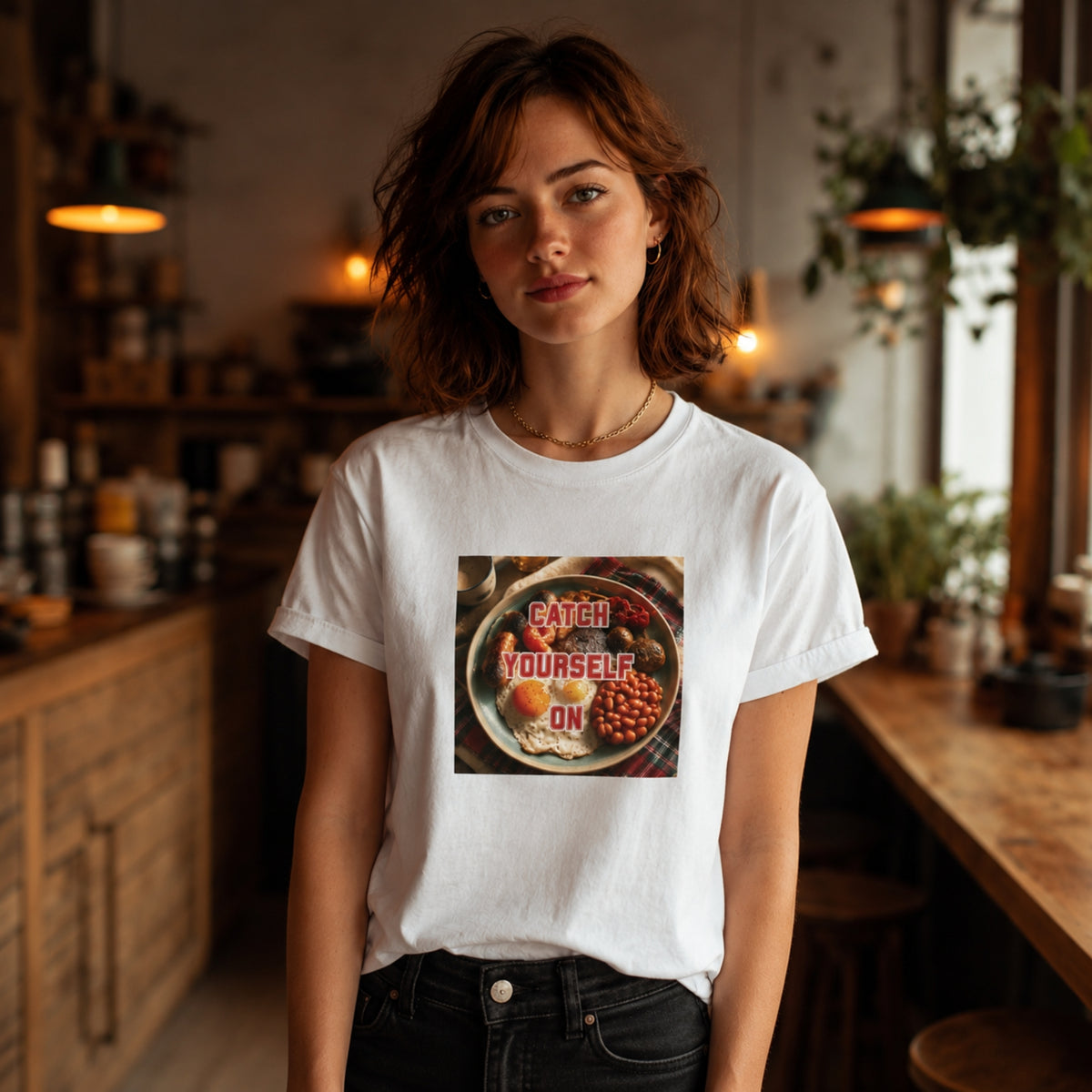 A woman with short brown hair stands in a warmly lit kitchen or cafe interior wearing a white graphic T-shirt. The shirt features a square print of a bowl with eggs and beans and the words 'CATCH YOURSELF ON' in red.
